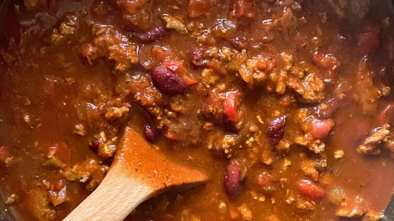 Overhead close-up of turkey chili with kidney beans, tomatoes, and red bell peppers in a pot with a wooden spoon.