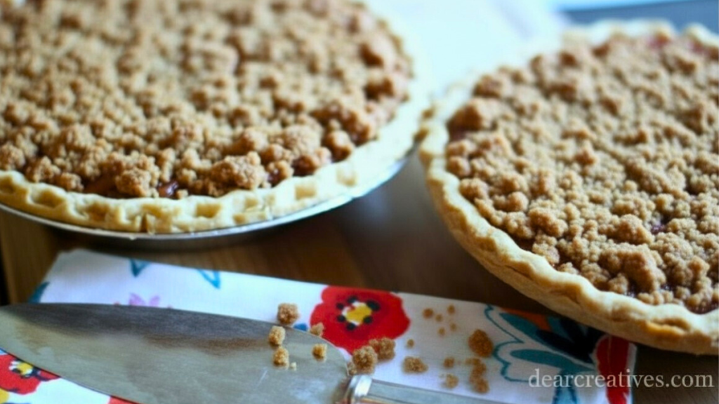 two peach pies with a crumb topping and next to a pie server on a napkin.
