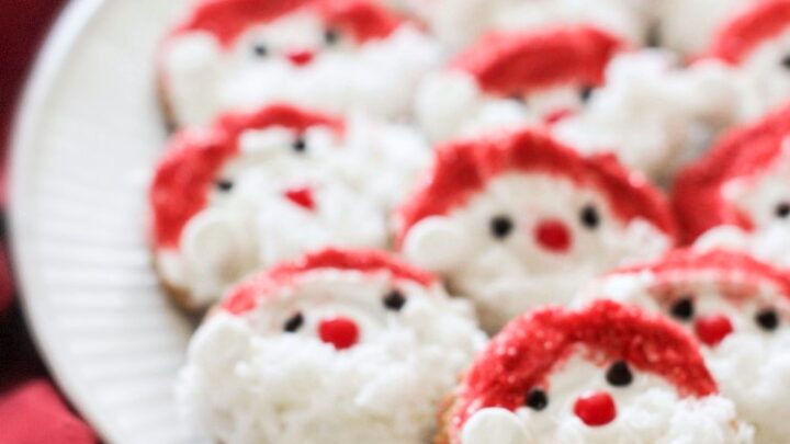 Finished Santa Claus cookies decorated with white icing then a simple red, white, sanding sugar a red hot cinnamon candy for the nose, and mini chocolate kisses for the eyes. Santa's beard is coconut flakes. And a mini marshmallow for on the Santa's red hat. These Christmas cookies are perfect for holiday baking with kids.