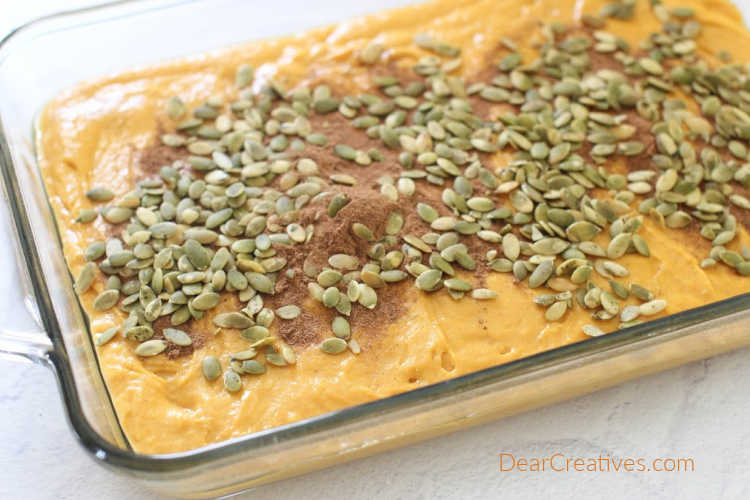 Pumpkin Cake Batter in a baking dish with pumpkin spice and pumpkin seeds on top ready to be swirled with a knife then baked. DearCreatives.com