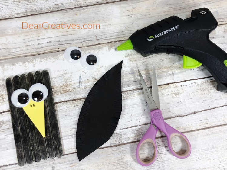 Step 5 - Embellish the Crow with two wiggle eyes, a large yellow craft foam triangle beak...See full instructions for making a crow, scarecrow and corn from craft sticks - DearCreatives.com