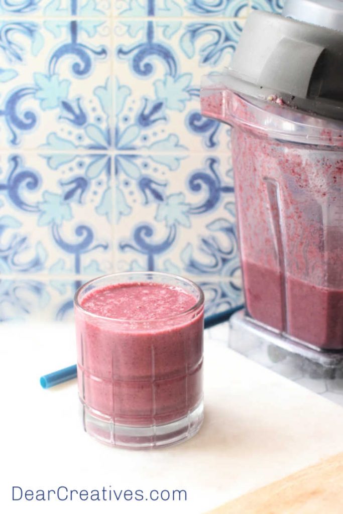 Blueberry-Smoothie-in-a-glass-next-to-a-Vitamix-blender-with-freshly-blended-smoothie-recipe-with-blueberries.-©-DearCreatives.com