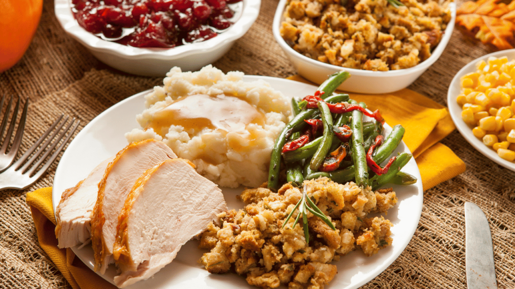 Closeup of a Turkey dinner with moist looking turkey slices, mashed potatoes and gravy, green beans and stuffing on a plate in the background cranberry sauce, stuffing and corn in serving bowls.