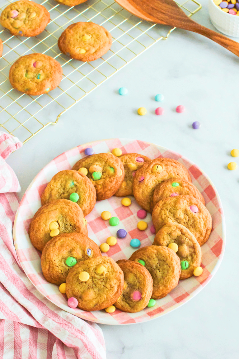 small batch M&M cookies on a plate and more cooling on a cookie rack in the background. a gingham kitchen towel by the warm plate of cookies, and a spatula on the bakers cooling rack and nearby a bowl of pastel M&Ms