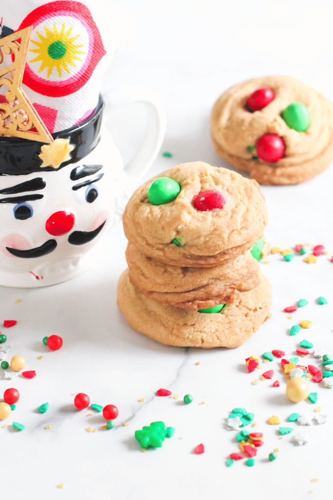 M&M Christmas Cookies with green and red M&Ms in them and stacked on a counter with festive Christmas sprinkles and a nutcracker cup and napkin.