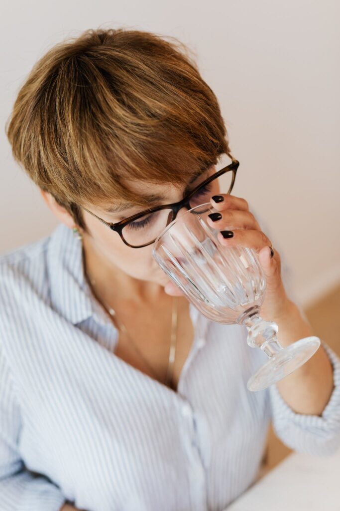 From above of female with short hair in eyeglasses and shirt with stripes drinking water while sitting at white table
