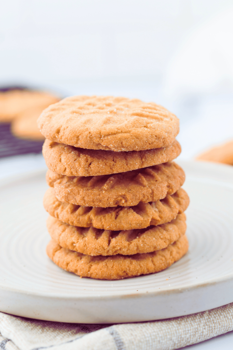 Peanut butter cookies stacked on a plate after cooled. With a baking rack with more cookies in the background.