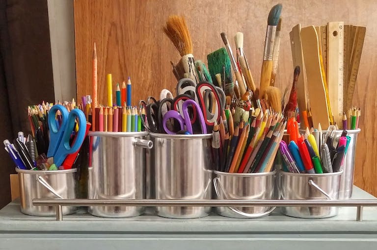 A diverse collection of art and craft supplies in metallic buckets on a wooden table.