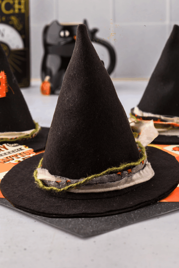 A witch hat made with felt with ribbons and twines wrapped around the brim. The brim has yet to be trimmed equally. In the background black cat cup, witchy decorations, and witch hats. The center witch hat brim will be trimmed to finish the felt craft project. DearCreatives.com