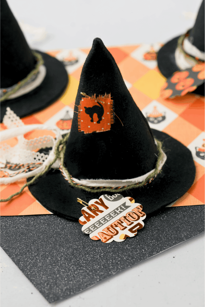 DIY Felt Witch Hat with a swatch of fabric with a scared cat pinned to it and ribbons and twine. The base of the hat has a cardstock cut out with eeeek scary caution and boo partially showing pinned to the witch hat brim. Adorable felt witch hats. DearCreatives.com