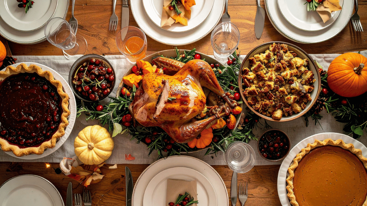 A festive Thanksgiving dinner table showing the centerpiece roast turkey, surrounded by side dishes including stuffing, cranberry sauce, and two homemade dessert pies.
