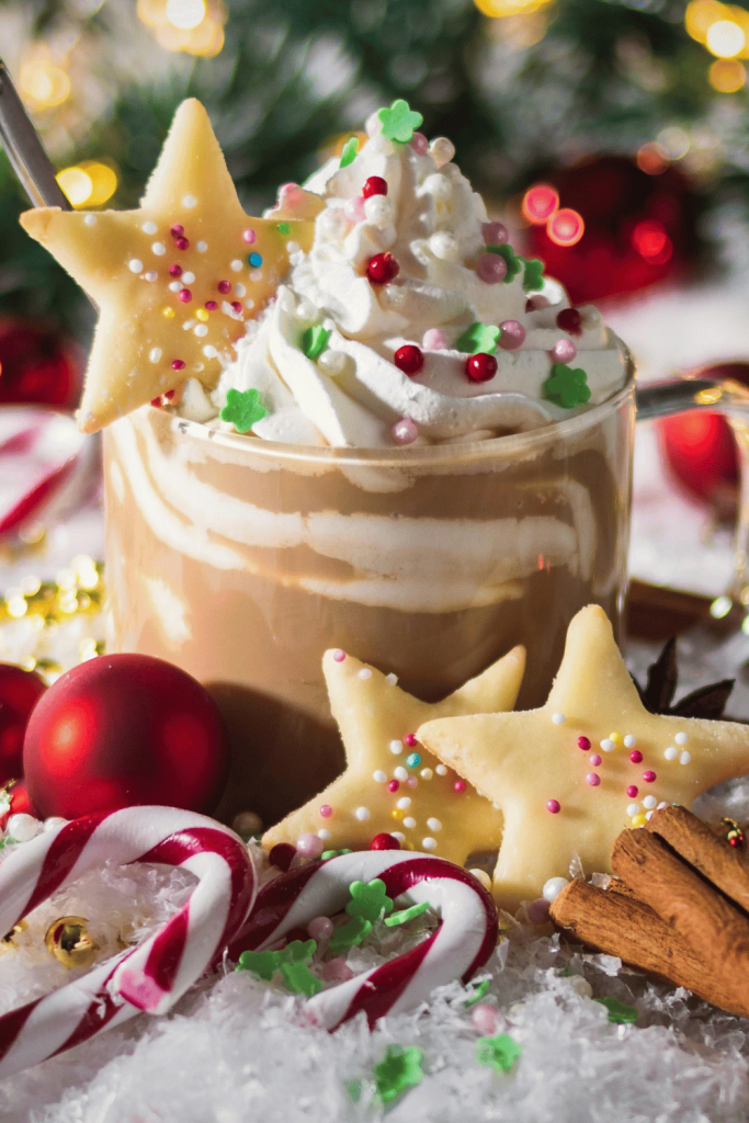 Festive hot chocolate with sprinkles and a cookie on top surrounded by cookies, candy canes and in the background festive holiday decorations