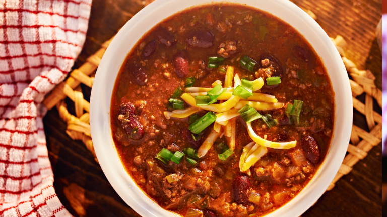 big bowl of chili with meat and beans close up topped with shredded cheddar cheese and chopped green onions.