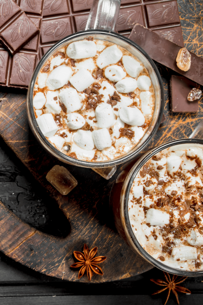 close up of two hot chocolates with chocolate with cocoa sprinkles and sitting on a wood  serving board with chocolate and chocolate with nuts. 
