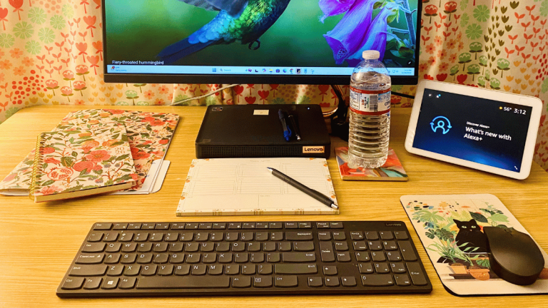 A bright, organized workspace featuring a computer monitor, a floral folder, and a bespoke floral planning calendar. A weekly agenda with a pen sits nearby, alongside an Amazon Echo Show and a glass of water on a coaster.