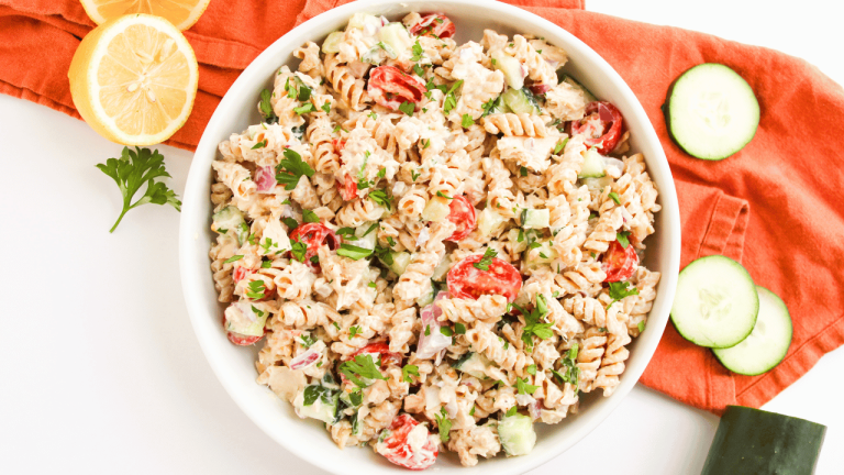 Healthy, Creamy, Tuna Pasta Salad in a bowl with light tuna, Greek yogurt, mayonnaise, chopped: cucumbers, tomatoes, and parsley. In the background a pretty linen under the bowl, sliced lemons, cucumbers, and parsley for garnishing.