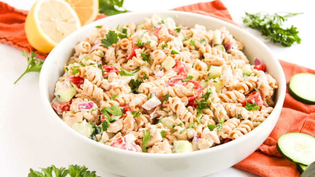 tuna pasta salad with vibrant vegetables, tomatoes, parsley, cucumbers, in a bowl ready to serve. In the background a bright linen, sliced lemons, parsley springs, and cucumber slices.