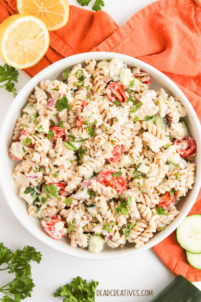 Make-Ahead Healthy, Creamy, Tuna Pasta Salad in a bowl setting on a linen, with lemons, parsley, and cucumbers in the background.