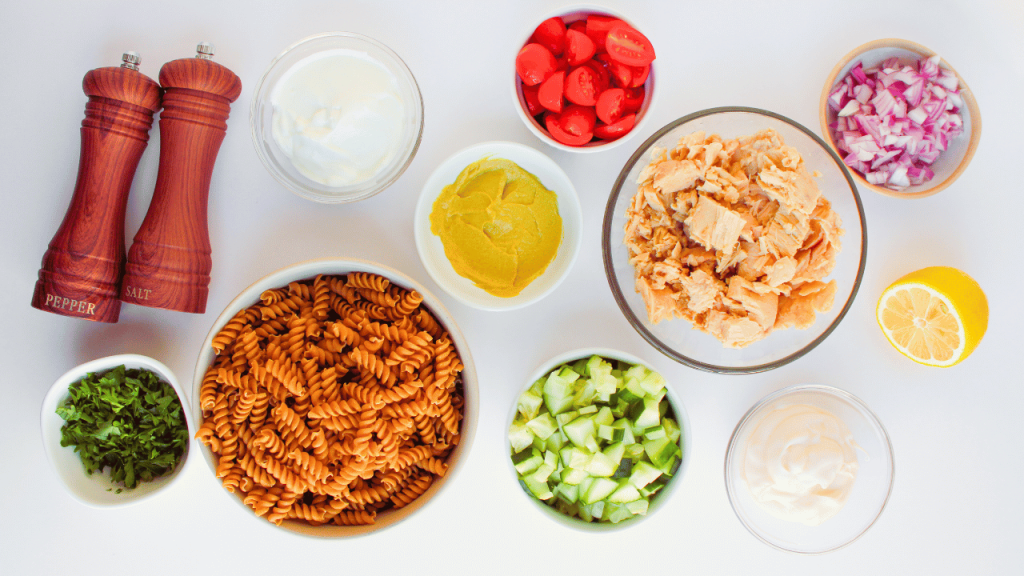 ingredients for a healthy tuna pasta salad pasta noodles, flakey tuna, chopped red onion, tomatoes, cucumbers, fresh parsley, and condiments.