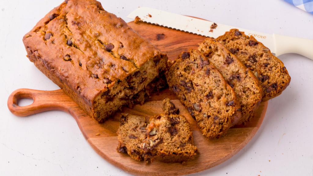 gluten-free banana bread with chocolate chips on a cutting board showing closeup of the loaf and slices stacked with a bread cutting knife sitting on a wood cutting board.
