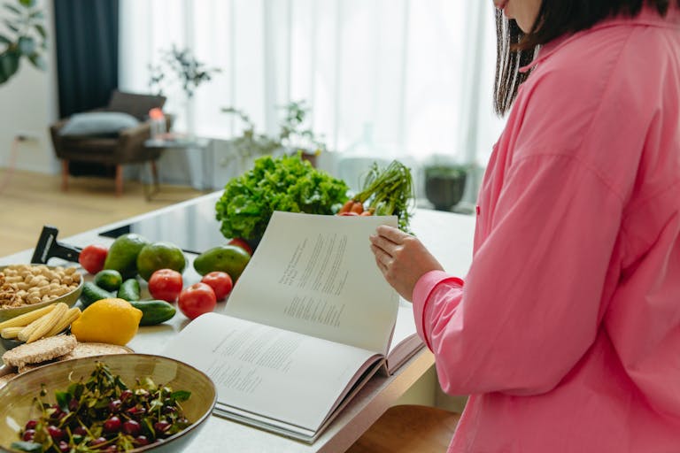 A woman in a kitchen flipping through a canning cookbook surrounded by fresh vegetables.