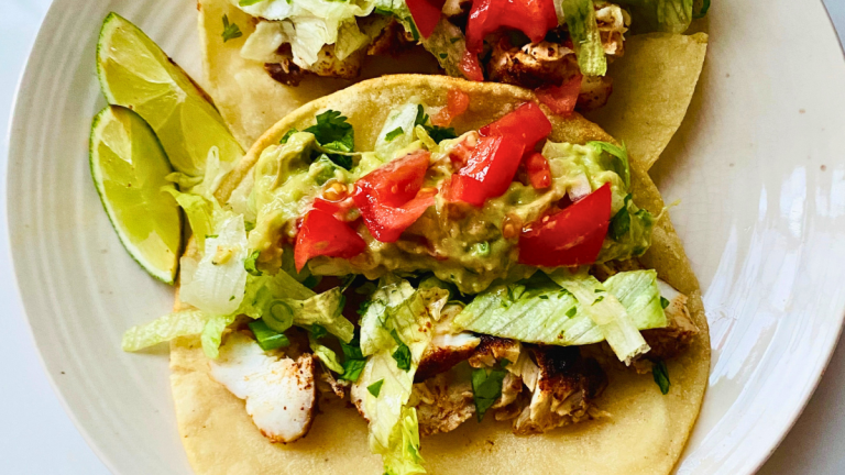 closeup image of two flavorful cod fish tacos on a plate with fresh homemade guacamole and taco toppings with two lime slices