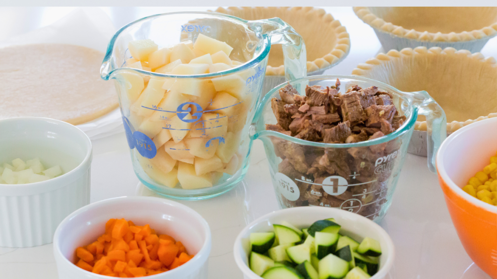 Beef pot pie ingredients with unbaked pastry shells in tins, pastry crust for topping, and chopped vegetables ready to make the filling.