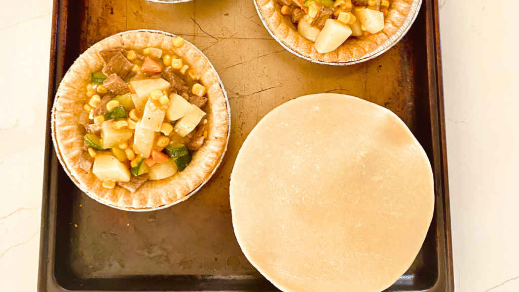Homemade beef pot pies being topped with round pastry crusts before crimping the edges and adding vents for baking.
