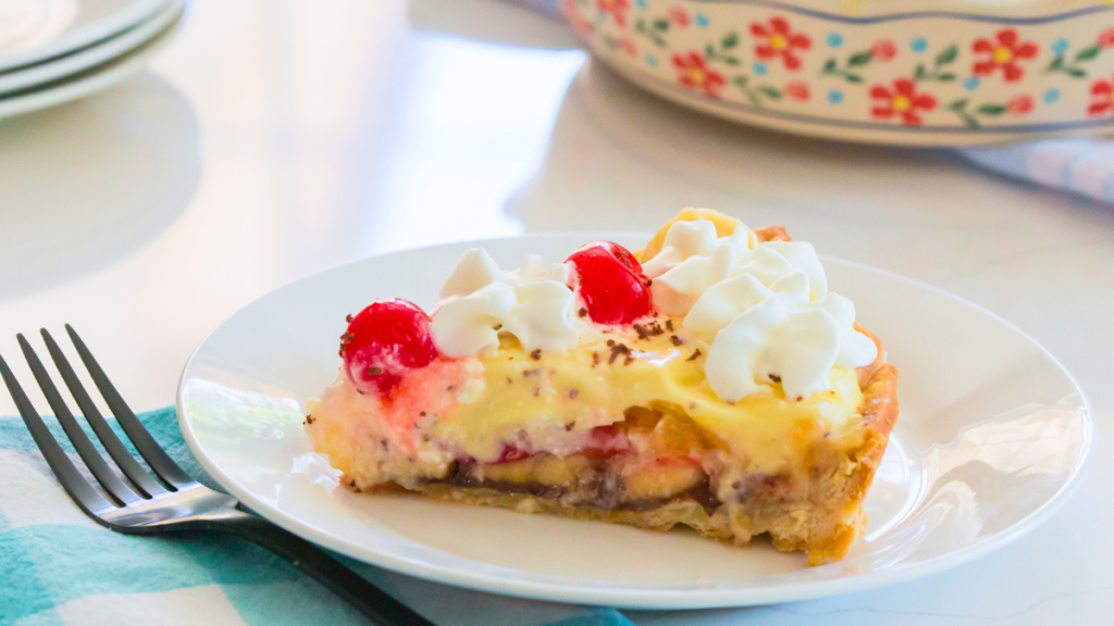 photo of banana split pie sliced in a pastry shell with layers of sliced fruit, sliced bananas, strawberries, and crushed pineapple on a layer of chocolate and garnished with whipped cream cherries with chocolate sprinkles